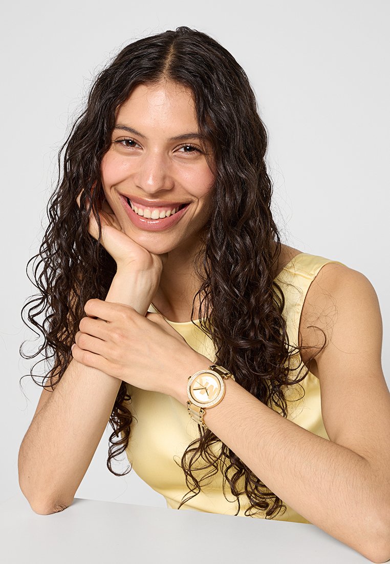 Montre-bracelet dorée avec un cadran rond, des accents en cristal et des index stylisés, portée avec un vêtement jaune clair.