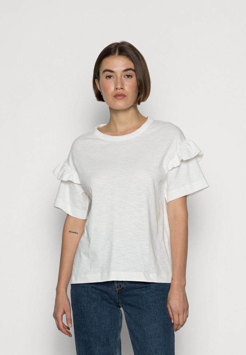 Hvit kortermet t-skjorte laget av mykt stoff med volanger på ermene. Rund hals og avslappet passform, kombinert med blå jeans.