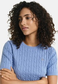 Pull en maille légère, bleu clair, à manches courtes, avec un col côtelé et un petit logo brodé sur la poitrine. Texturé et doux.