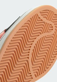 Suola della scarpa in gomma arancione con motivo a zigzag. Il bordo presenta una linea bianca liscia, in contrasto con i materiali superiori neri e grigi.