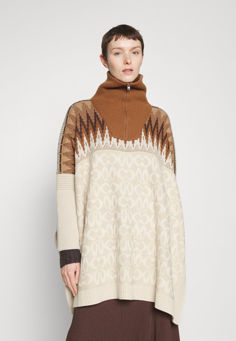 Person mit kurzen Haaren, die einen hochgeschlossenen beigen und braun gemusterten Oversize-Pullover und einen braunen gerippten Rock trägt, steht vor einem schlichten Hintergrund.