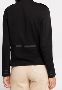 Zwarte getailleerde jas met een gladde textuur, voorzien van schouderepauletten en satijnen accenten op de mouwen, gedragen over beige broek.