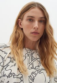 Top bianco con motivi astratti neri, caratterizzato da un design plissettato e un colletto alto, tessuto testurizzato e dettagli sulle spalle.