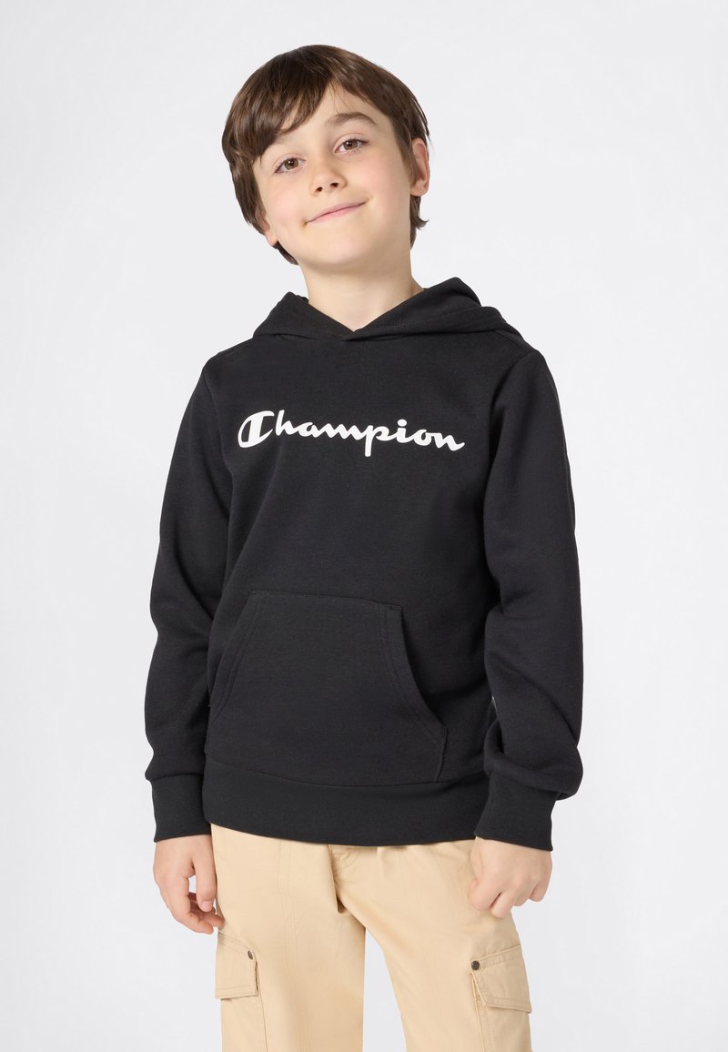 Ragazzo sorridente che indossa un felpa nera Champion e pantaloni cargo beige, in piedi contro uno sfondo chiaro e semplice.