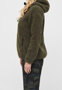 Olivgrön fleece-hoodie med en strukturerad yta, justerbar huva och sidofickor med dragkedja, kombinerad med m örka camouflagemönstrade cargobyxor.