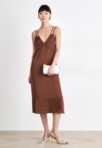 Rochie midi texturată, de culoare maro, cu bretele subțiri și decolteu în V. Se ține o poșetă albă cu accente aurii, asortată cu pantofi cu toc metalic.