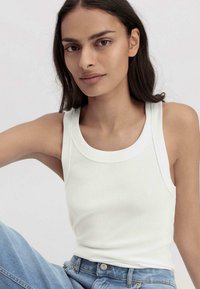 Weißes, geripptes Tanktop mit rundem Halsausschnitt und schmalen Trägern, kombiniert mit blauen Jeans. Glatte Textur und taillierte Passform.