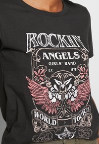Černé bavlněné tričko s vintage designem rockové kapely, bíl�ými a růžovými grafikami zahrnujícími křídla, květiny a text o turné.