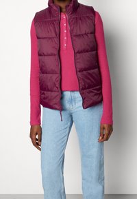 Burgundy pufferväst med hög krage, dragkedja och quiltat mönster, lager på lager över en rosa långärmad topp och ljusblå jeans.