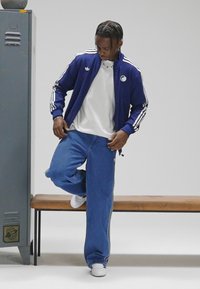 Μπλε αθλητικό μπουφάν με φερμουάρ και λευκές ρίγες, φορεμένο πάνω από λευκό πουκάμισο και φαρδιά μπλε τζιν, σετ με λευκά αθλητικά παπούτσια, σε παγκάκι.