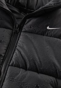 Μαύρο μπουφάν τύπου puffer με γυαλιστερό φινίρισμα, με φερμουάρ στο μπροστινό μέρος και σταγόνες νερού στην επιφάνεια. Το λογότυπο της Nike είναι ορατό στο στήθος.