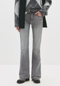 Osoba v šedých rozšírených džínach, čiernych topánkach a šedom svetri so vzorom, s jednou rukou vo vrecku na bielom pozadí.