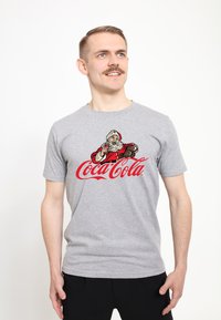 Muž v šedém tričku s vintage Santou, který drží láhev Coca-Coly nad červeným logem Coca-Cola, stojící na bílém pozadí.