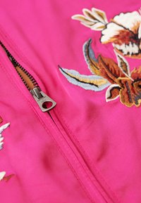 Veste fuchsia avec une broderie florale complexe en marron, bleu et ivoire. Présente une fermeture éclair en métal argenté sur le devant. Texture de tissu lisse.