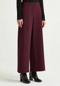 Pantaloni a gamba larga in tessuto color bordeaux, con vita alta e texture liscia. Abbinati a tacchi neri con motivo testurizzato.