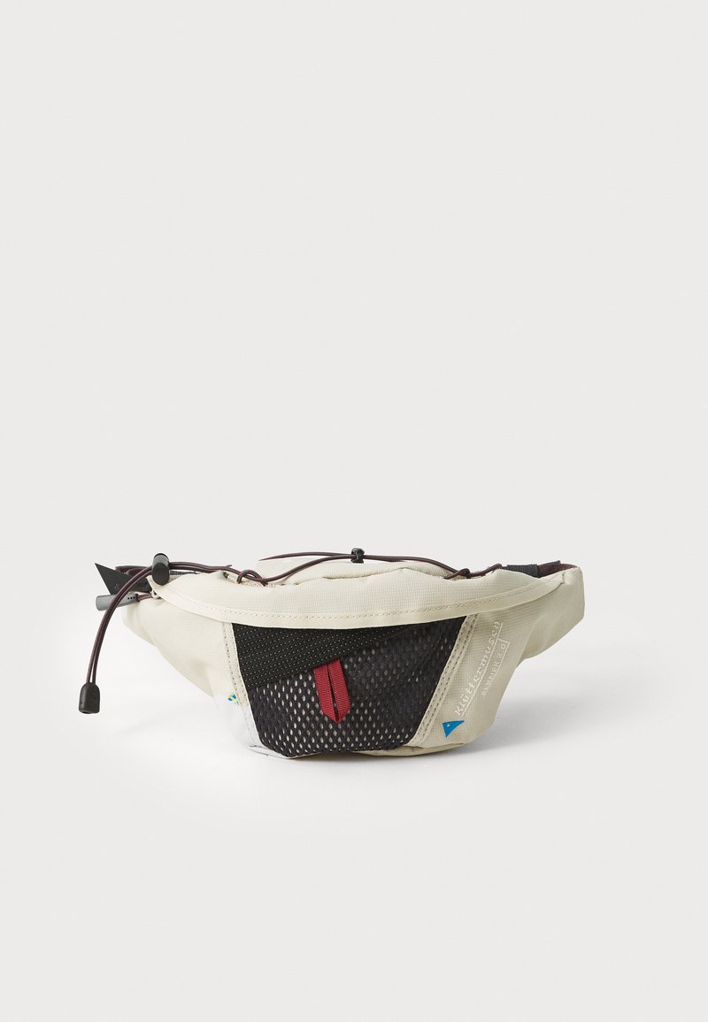 Kremi spalvos fanny pack su juodu tinkleliu, raudonu akcentu, reguliuojamu virvute ir logotipo prekės ženklo žymė; kompaktiškas, lenktas dizainas, užtikrinantis universalumą.