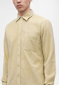 Gros plan sur une personne portant une chemise en velours côtelé beige à boutons avec une poche poitrine côté gauche.