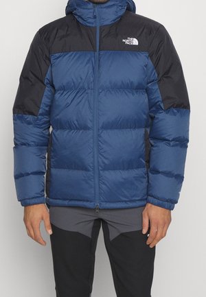 Mężczyzna noszący niebiesko-czarną puchową kurtkę z logo The North Face na piersi, w połączeniu z ciemnymi spodniami.