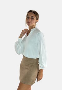 Blouse blanche texturée avec un col mandarin et des manches froncées, associée à une mini jupe en daim beige avec une fermeture éclair à l'arrière.