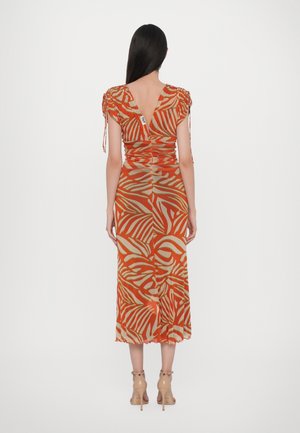 Woman with long dark hair wearing an orange and beige patterned midi dress with short sleeves and beige high-heeled sandals, standing facing away.