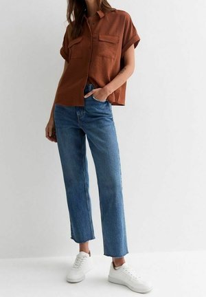 Kvinde iført rustfarvet kortærmet skjorte med knapper, blå korte jeans og hvide sneakers, stående mod en helt hvid baggrund.