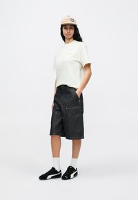 Joven con gorra beige, camiseta de color crudo, pantalones cortos largos de mezclilla negra, calcetines blancos y zapatillas Puma negras, de pie con las manos en los bolsillos.