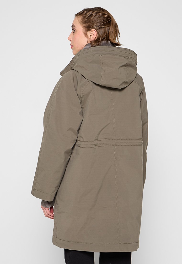 Frau in einer overzised olivgrünen Kapuzenparka, von hinten gezeigt, mit entspannten Händen an den Seiten.