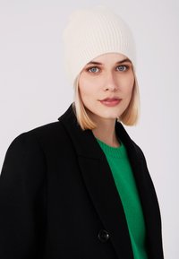 Gorro blanco acanalado hecho de un material suave, con un ajuste ceñido. Llevado con un abrigo negro y un suéter verde. Fondo neutro.