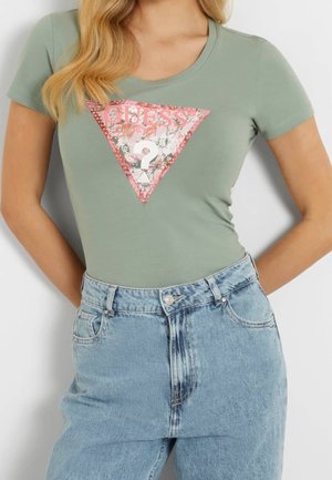 Kvinde iført lysegrøn tætsiddende t-shirt med lyserødt blomstret Guess-logo i trekant og lyseblå højtaljede jeans.