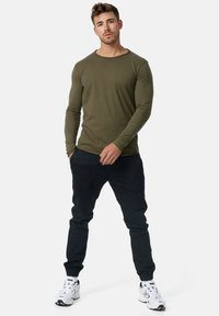 Olivengrøn langærmet skjorte parret med mørke joggerbukser og hvide sneakers. Skjorten har en rund udringning og tætsiddende ærmer; bukserne har sidelommer.