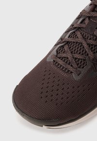 Chaussure de sport marron foncé avec une tige en maille texturée, un embout renforcé et un design de lacet complexe, avec des accents gris et des trous de ventilation.