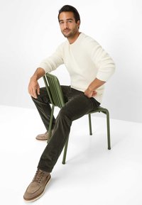 Beige gebreide trui, donker groene corduroy broek en lichtbruine suède schoenen. Zittend op een groene metalen stoel tegen een witte achtergrond.