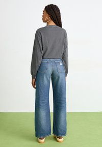 Woman with long braided hair wearing hoop earrings, gray long-sleeve shirt, wide-leg blue jeans, and beige sneakers standing on green floor.