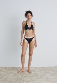 Juodas bikini rinkinys su trikampiu viršumi ir žemu sijonu, su baltu logotipo tekstu. Modelis stovi ant tekstūruoto šviesaus akmens grindų.