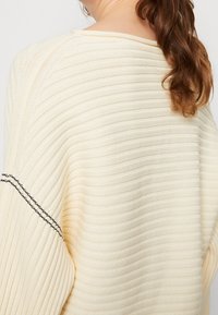 Cream ribbed knit sweater with a loose fit. Features a wide neckline and black stitching accents on the sleeves. Textured fabric.