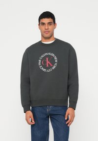 Tmavosivá bavlnená mikina s okrúhlym logom s nápisom "CALVIN KLEIN NEW YORK" a "1968" v bielom a červenom písme. Neformálny strih s rebrovanými manžetami.