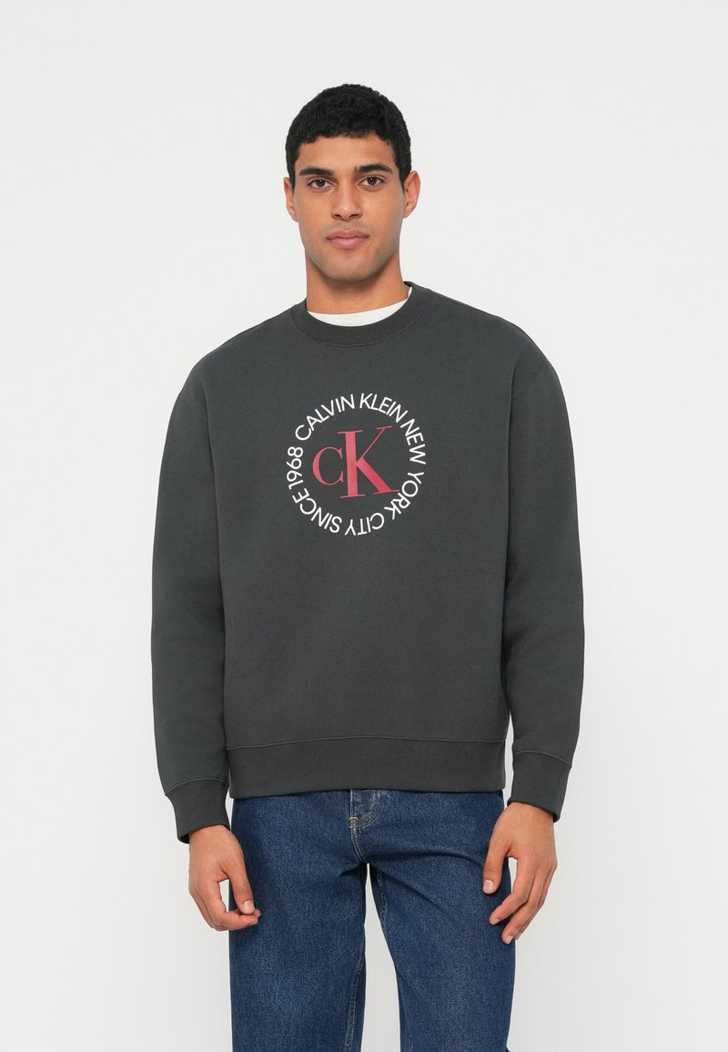 Tmavosivá bavlnená mikina s okrúhlym logom s nápisom "CALVIN KLEIN NEW YORK" a "1968" v bielom a červenom písme. Neformálny strih s rebrovanými manžetami.