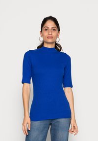 Enganliegendes, blaues Rippoberteil mit hohem Kragen und kurzen Ärmeln. Der Stoff ist glatt. Kombiniert mit blauen Jeans.