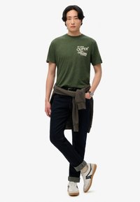 Grøn heathered t-shirt med hvidt logo, mørke jeans og hvide sneakers med brune accenter. En sweater bundet omkring taljen giver kontrast.