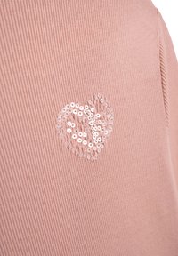 Rosa gerippter Stoff mit einer herzförmigen Applikation aus weißen Pailletten auf der linken Seite. Die Struktur erscheint aufgrund der Pailletten leicht erhaben.