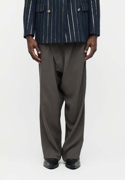 Χέρια στα πλάγια που φορούν σκούρο καφέ παντελόνι με φαρδύ πόδι, μαύρα παπούτσια και μπλε σκούρο σακάκι με λεπτές χρυσές κάθετες ρίγες και χρυσά κουμπιά.