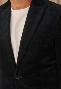 Navy corduroy blazer with a notched lapel, single button closure, and a chest pocket. Worn over a white T-shirt. Textured fabric.