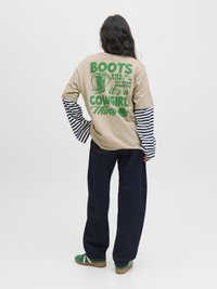 Beige übergroßes T-Shirt mit grünem Grafiktext auf dem Rücken, gestreiften langen Ärmeln darunter, kombiniert mit dunkelblauen weit geschnittenen Hosen und grünen Schuhen.