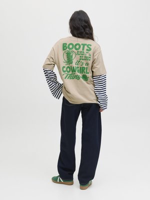 Beige oversized t-skjorte med grønn grafisk tekst på baksiden, stripete lange ermer under, kombinert med mørkeblå vide bukser og grønne sko.