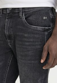 Black denim jeans feature a slight fade, five-pocket design, and a small metal logo on the front pocket. The fabric has a smooth texture.
