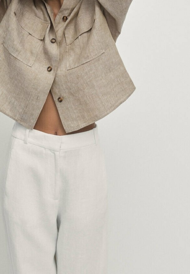 Personne portant une veste courte beige boutonnée avec de grandes poches et un pantalon blanc taille haute, les bras levés au-dessus de la tête sur un fond uni.