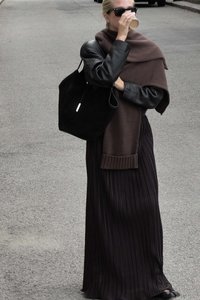 Pitkä laskostettu tumma hame, musta nahkatakki, ruskea neulehuivi, aurinkolasit, kahvikuppi kädessä, musta laukkukassi, kaupunkimaisema.