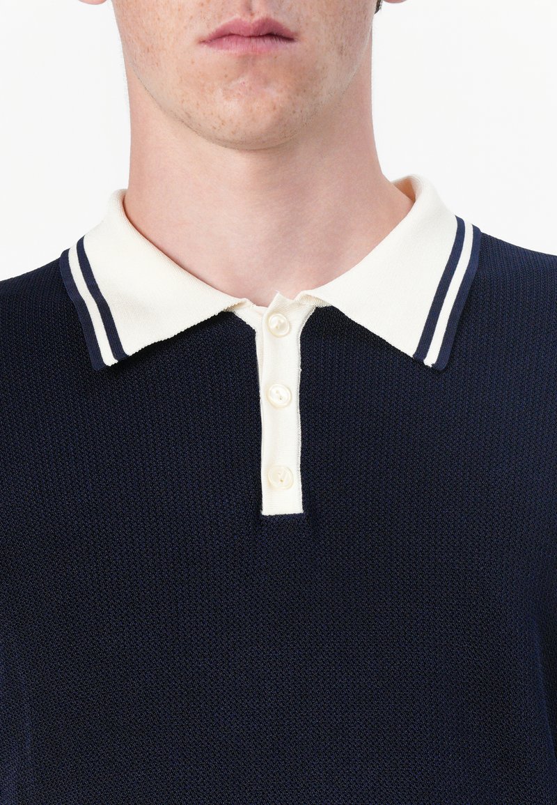 Marineblaues Strick-Polohemd mit einem kontrastierenden krämfarbenen Kragen, der marineblaue Streifen hat, und einer Knopfleiste mit vier Knöpfen. Nahaufnahme des strukturierten Stoffs.