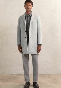 Helehall villane mantli, millel on sakiline krae, ühe nööbiga disain ja mustad nööbid, koos hallide pükstega ja mustade saapatega.