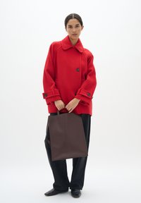 Rode wolmix oversized jas met grote knopen en manchetdetails, gecombineerd met een eenvoudige bruine totebag en donkere broeken. Zwarte schoenen maken de look compleet.